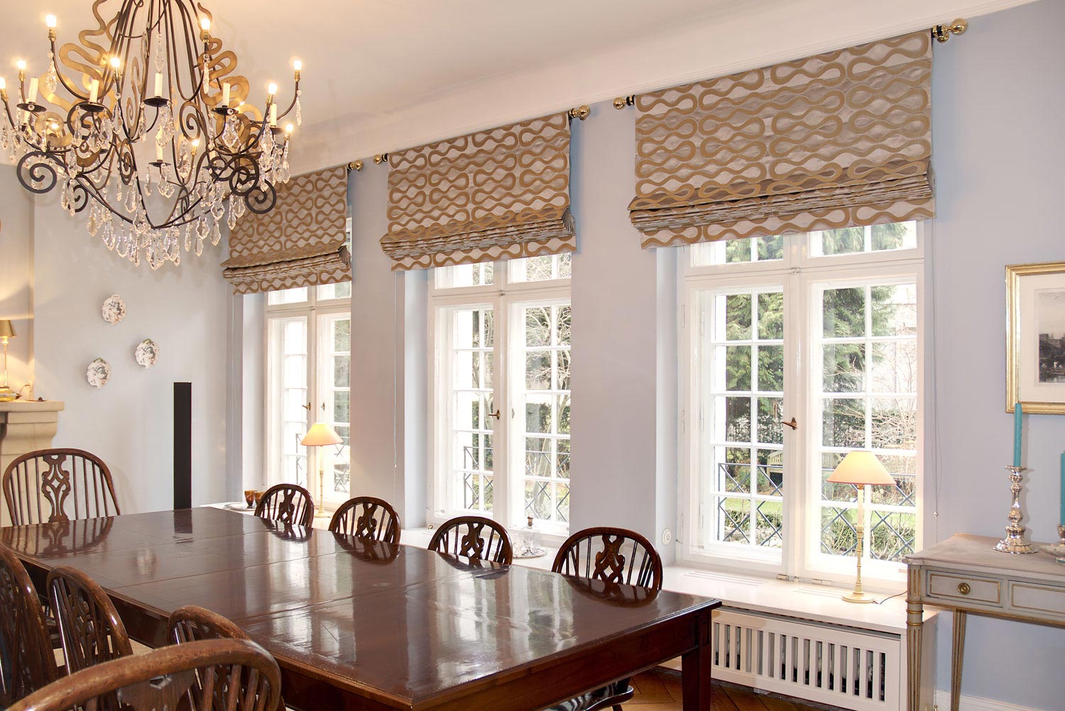 Elegantes Esszimmer mit Faltrollos mit geradem Rolloabschluss in gold-beigem Muster und dekorativen Stilstangen vor großen Sprossenfenstern.